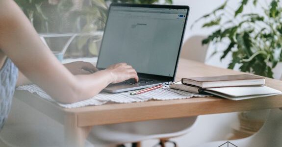 Eine Person sitzt an einem Schreibtisch, man sieht nur den Ausschnitt des Schreibtischs und der Arme und Hände der Person, wie sie in einen Laptop tippen, Work Situation