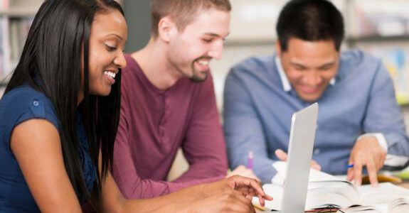 Drei Personen sitzen an einem Schreibtisch und arbeiten mit Laptop und Notizen