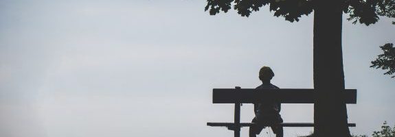 Eine Person sitzt alleine und mit dem Rücken zu uns gekehrt auf einer Bank unter einem Baum