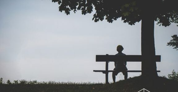 Eine Person sitzt alleine und mit dem Rücken zu uns gekehrt auf einer Bank unter einem Baum