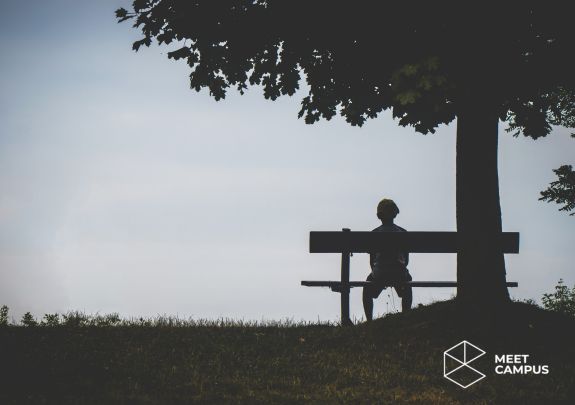 Eine Person sitzt alleine und mit dem Rücken zu uns gekehrt auf einer Bank unter einem Baum