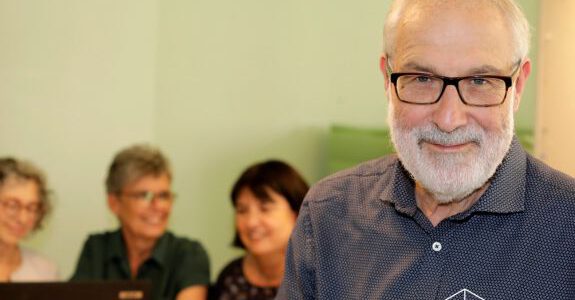 Mann mit Brille und Hemd im Fokus vorne rechts schaut in die Kamera, im Hintergrund verschwommen sitzen drei Personen vor einem Laptop und arbeiten gemeinsam