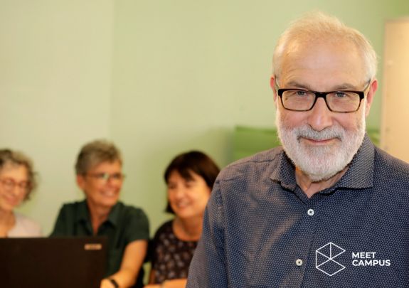 Mann mit Brille und Hemd im Fokus vorne rechts schaut in die Kamera, im Hintergrund verschwommen sitzen drei Personen vor einem Laptop und arbeiten gemeinsam